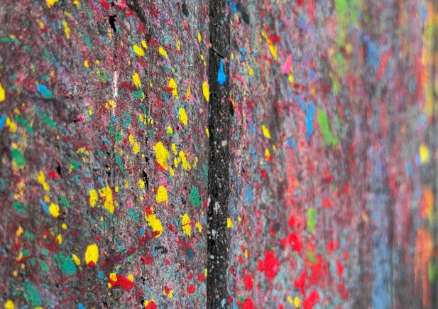 Nahaufnahme einer Betonwand, die mit leuchtenden Farben, darunter Gelb, Rot, Grün und Blau, bespritzt ist, was ihr ein lebendiges, abstraktes Aussehen verleiht. Eine vertikale Linie unterteilt die Wand in zwei Abschnitte.