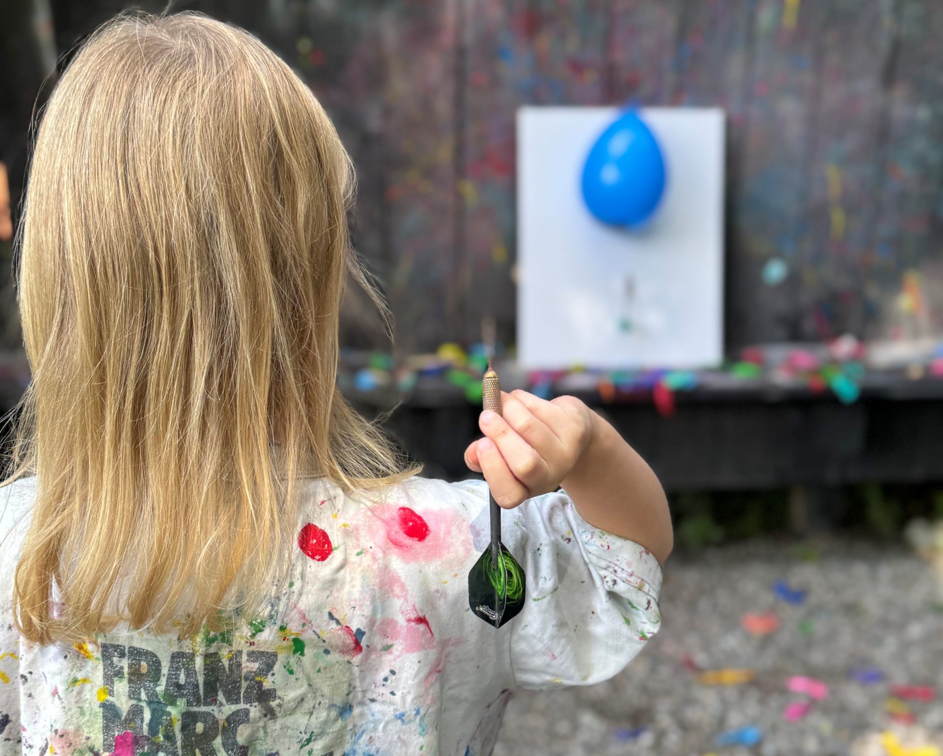 Ein Kind mit langen blonden Haaren, das ein mit Farbe bespritztes Hemd trägt, steht im Freien und hält einen Dartpfeil in der Hand. Es steht vor einer an der Wand befestigten Leinwand mit einem blauen Luftballon und bereitet sich darauf vor, den Pfeil zu werfen.