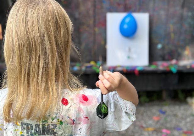 Ein Kind mit langen blonden Haaren, das ein mit Farbe bespritztes Hemd trägt, steht im Freien und hält einen Dartpfeil in der Hand. Es steht vor einer an der Wand befestigten Leinwand mit einem blauen Luftballon und bereitet sich darauf vor, den Pfeil zu werfen.