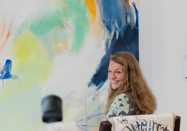 Eine Frau mit langen Haaren lächelt in die Kamera, während sie vor einem bunten Action Painting sitzt. Im Vordergrund richtet jemand eine Kamera auf sie. Auf der Rückenlehne ihres Stuhls befindet sich eine Weste mit der Aufschrift NICHTS TUN.