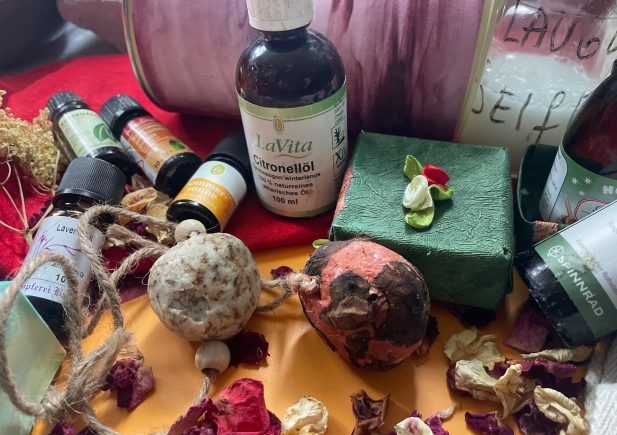 Eine Sammlung von Flaschen mit ätherischen Ölen aus der Seifenwerkstatt, handgefertigten Seifen, getrockneten Blumen und dekorativen Badeartikeln, die auf einer Fläche mit farbenfrohen Verpackungen und natürlichen Texturen angeordnet sind.