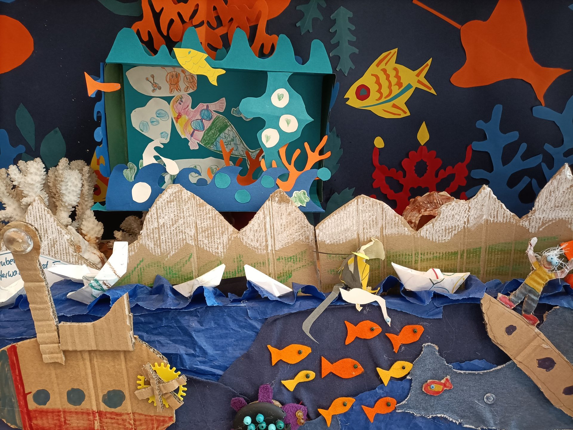 Ein farbenfrohes Unterwasser-Diorama mit Schiffen aus Pappe, Papierbergen, Korallen, Fischen und Meeresbewohnern. Orangefarbene Fische schwimmen in blauem Wasser, mit lebhaften Meeresbewohnern, Papierbooten und Pappkulissen in einer lebendigen Ozeanszene.