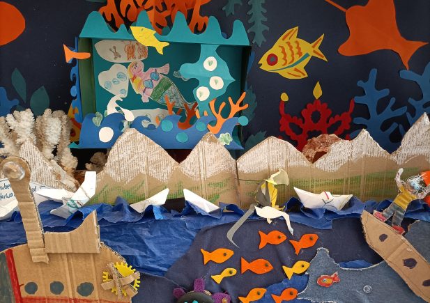 Ein farbenfrohes Unterwasser-Diorama mit Schiffen aus Pappe, Papierbergen, Korallen, Fischen und Meeresbewohnern. Orangefarbene Fische schwimmen in blauem Wasser, mit lebhaften Meeresbewohnern, Papierbooten und Pappkulissen in einer lebendigen Ozeanszene.