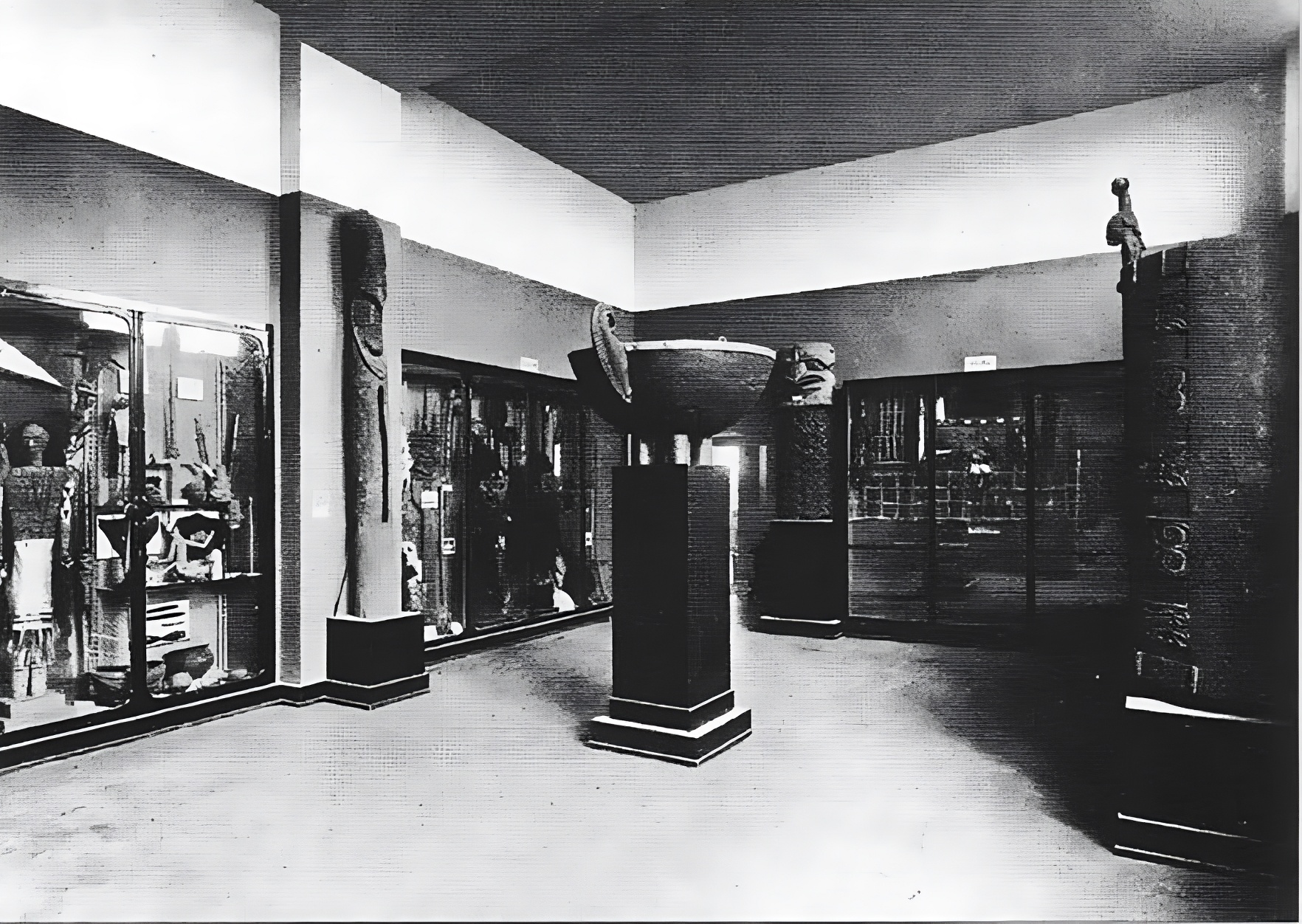 Ein Schwarz-Weiß-Foto einer Museumsgalerie zeigt große Skulpturen und Artefakte in Vitrinen und auf Sockeln, mit hohen Decken und reichlich Beleuchtung.