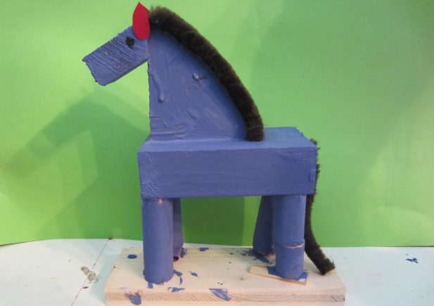 Eine handgefertigte Pferdefigur aus blau bemalter Pappe und Holzklötzen steht auf einem Holzsockel. Mähne und Schweif sind aus schwarzem Pfeifenreiniger, die Ohren aus rotem Filz, der Hintergrund ist grün.