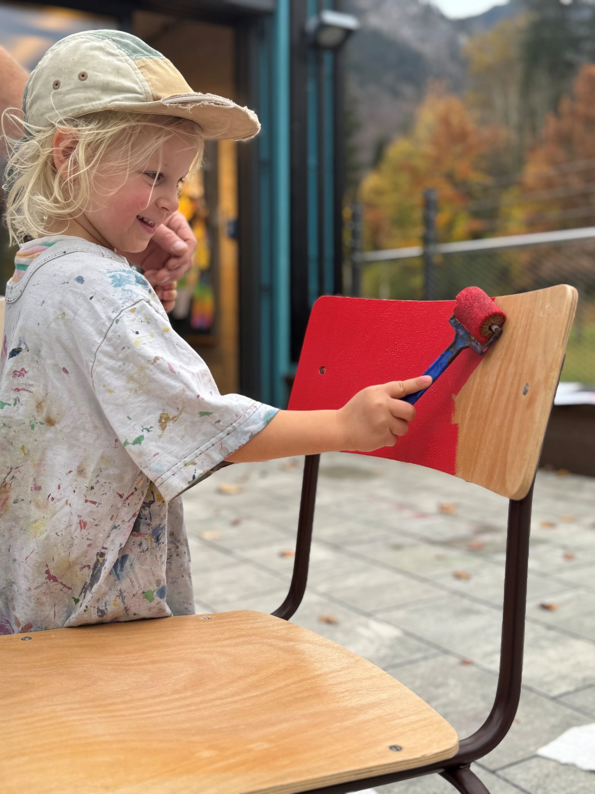 Ein lächelndes Kind, das ein mit Farbe bespritztes Hemd und eine Mütze trägt, bestreicht mit einem Farbroller die Rückenlehne eines Holzstuhls mit leuchtend roter Farbe. Im unscharfen Hintergrund sind Herbstbäume und ein Haus zu sehen.