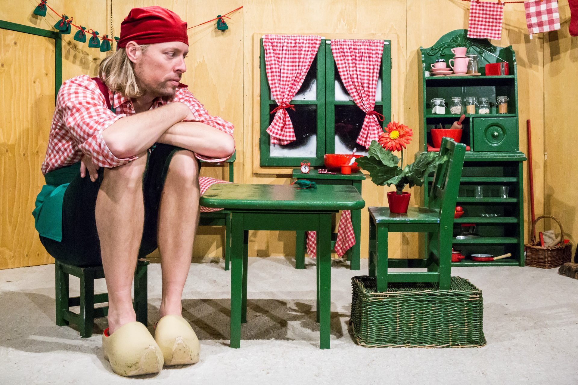 Ein Mann mit rotem Kopftuch und kariertem Hemd sitzt auf einem winzigen grünen Stuhl an einem kleinen Tisch in einer Miniaturküche und schaut nachdenklich, als ob er auf den Vortrag Geschichte der Zoos von Rasem Baban wartet. Die kindliche Einrichtung kontrastiert mit seiner übergroßen Präsenz.