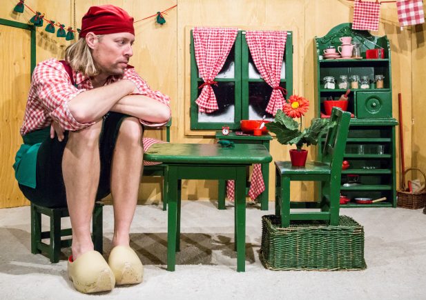 Ein Mann mit rotem Kopftuch und kariertem Hemd sitzt auf einem winzigen grünen Stuhl an einem kleinen Tisch in einer Miniaturküche und schaut nachdenklich, als ob er auf den Vortrag Geschichte der Zoos von Rasem Baban wartet. Die kindliche Einrichtung kontrastiert mit seiner übergroßen Präsenz.