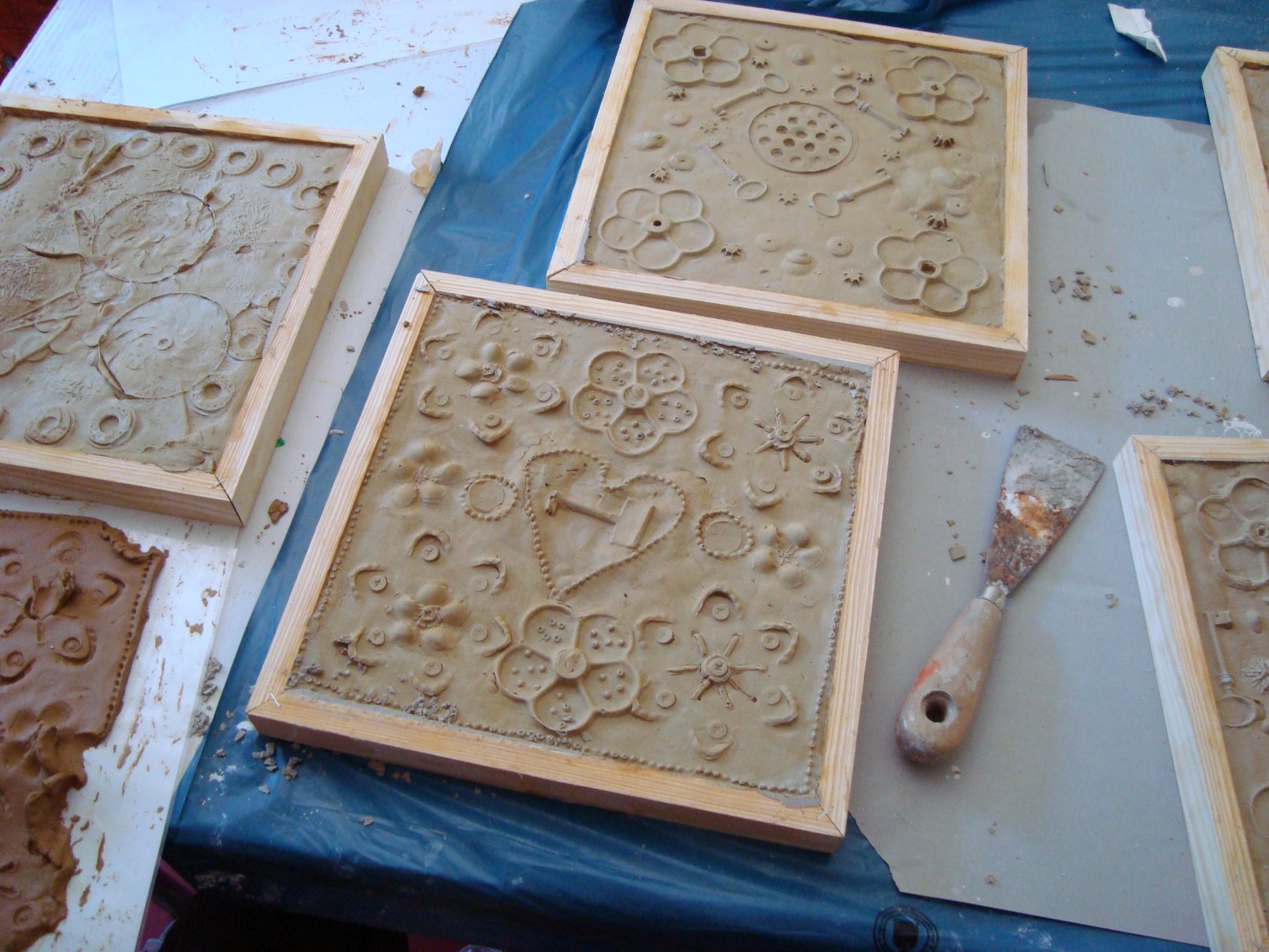 Tonfliesen mit dekorativen Mustern und Blumenaufdrucken sind in Holz gerahmt und auf einem Tisch platziert. Ein Metallspachtel liegt in der Nähe, was darauf hindeutet, dass die Fliesen Teil eines Kunst- oder Handwerksprojekts sind.