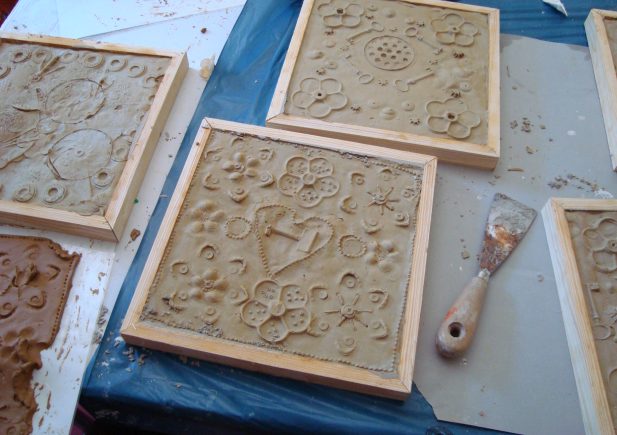 Tonfliesen mit dekorativen Mustern und Blumenaufdrucken sind in Holz gerahmt und auf einem Tisch platziert. Ein Metallspachtel liegt in der Nähe, was darauf hindeutet, dass die Fliesen Teil eines Kunst- oder Handwerksprojekts sind.