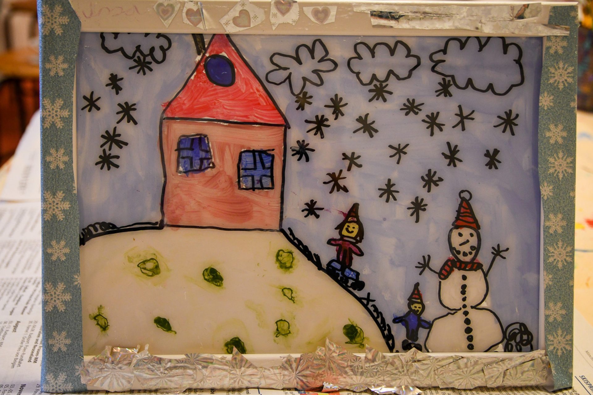 Die Zeichnung eines Kindes zeigt ein Haus mit rotem Dach auf einem verschneiten Hügel, zwei Kinder, einen Schneemann mit Hut und einen Himmel voller schwarzer Schneeflocken. Das Bild ist mit Silberfolie und Herzverzierungen eingerahmt.