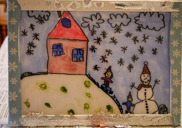 Die Zeichnung eines Kindes zeigt ein Haus mit rotem Dach auf einem verschneiten Hügel, zwei Kinder, einen Schneemann mit Hut und einen Himmel voller schwarzer Schneeflocken. Das Bild ist mit Silberfolie und Herzverzierungen eingerahmt.
