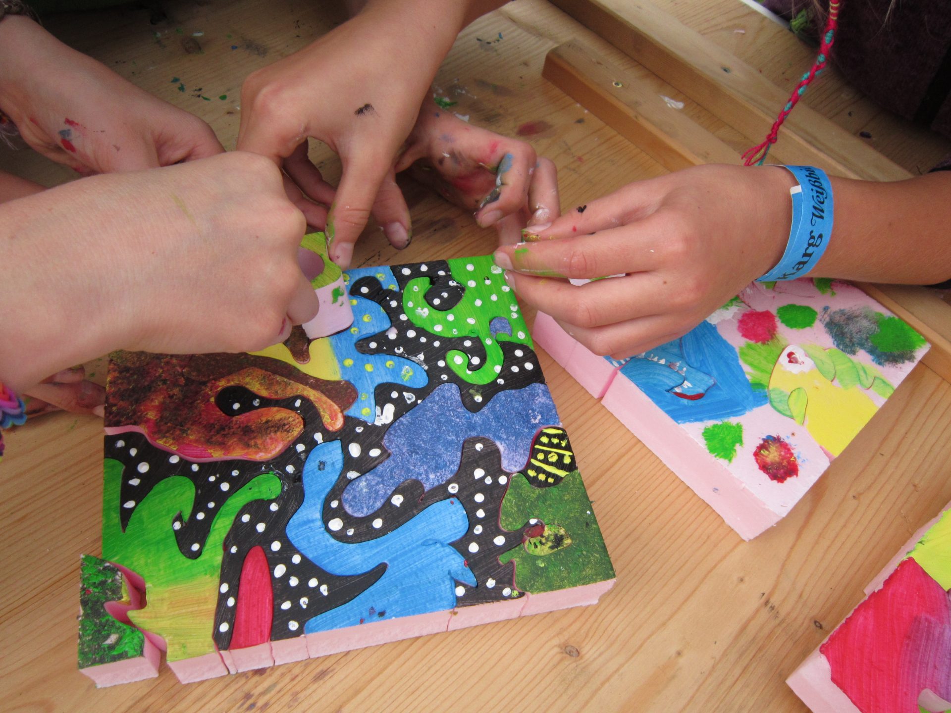Vier Hände arbeiten zusammen, um ein buntes, abstrakt bemaltes Puzzle mit Tier- und Naturformen auf einem Holztisch zusammenzusetzen. Einige Finger sind bemalt, und an einem Handgelenk ist ein blaues Festivalarmband zu sehen.