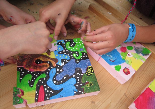 Vier Hände arbeiten zusammen, um ein buntes, abstrakt bemaltes Puzzle mit Tier- und Naturformen auf einem Holztisch zusammenzusetzen. Einige Finger sind bemalt, und an einem Handgelenk ist ein blaues Festivalarmband zu sehen.