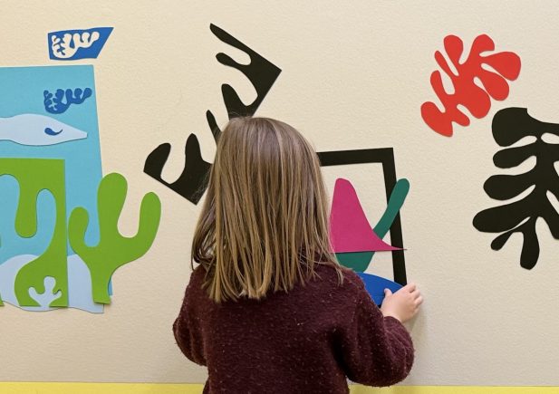 Ein junges Mädchen mit hellbraunem Haar steht vor einer Wand, die mit bunten, abstrakten ausgeschnittenen Formen verziert ist. Sie trägt ein kastanienbraunes Kleid, beige Leggings und grüne Stiefel. Der untere Teil der Wand ist gelb gestrichen.