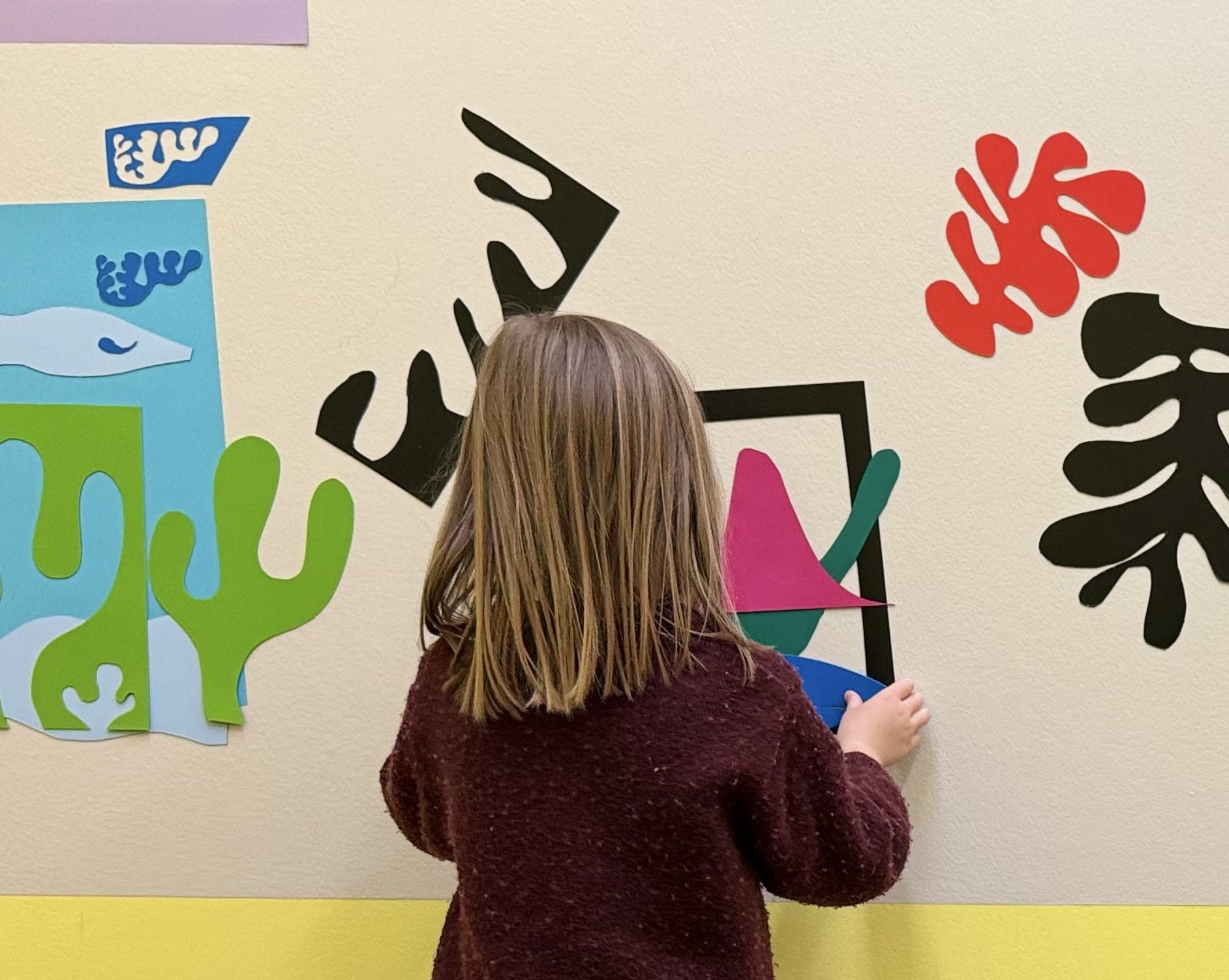 Ein junges Mädchen mit hellbraunem Haar steht vor einer Wand, die mit bunten, abstrakten ausgeschnittenen Formen verziert ist. Sie trägt ein kastanienbraunes Kleid, beige Leggings und grüne Stiefel. Der untere Teil der Wand ist gelb gestrichen.