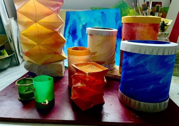 Bunte handgefertigte Papierlaternen in verschiedenen Formen und Größen sind auf einem Tisch ausgestellt, mit Kunstzubehör und hellen Gemälden im Hintergrund. Die Laternen sind orange, rot, grün, blau und gelb.