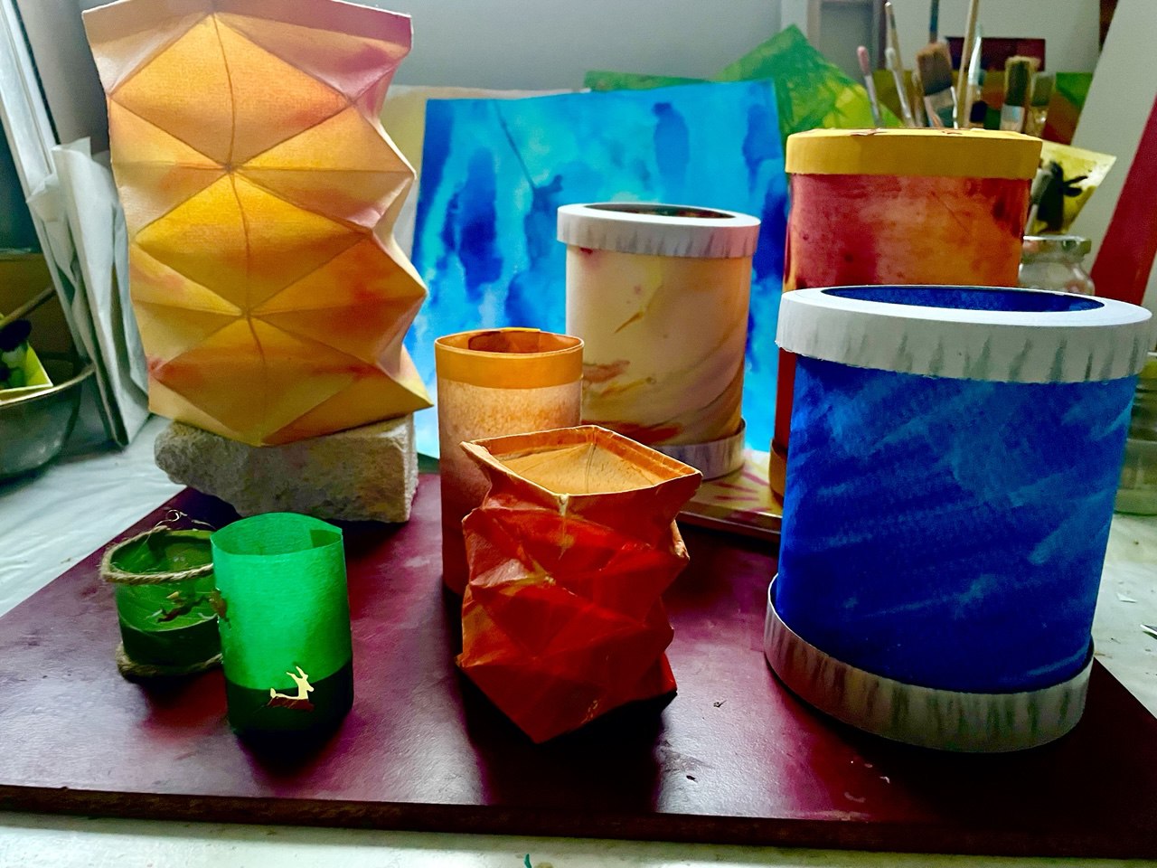 Bunte handgefertigte Papierlaternen in verschiedenen Formen und Größen sind auf einem Tisch ausgestellt, mit Kunstzubehör und hellen Gemälden im Hintergrund. Die Laternen sind orange, rot, grün, blau und gelb.