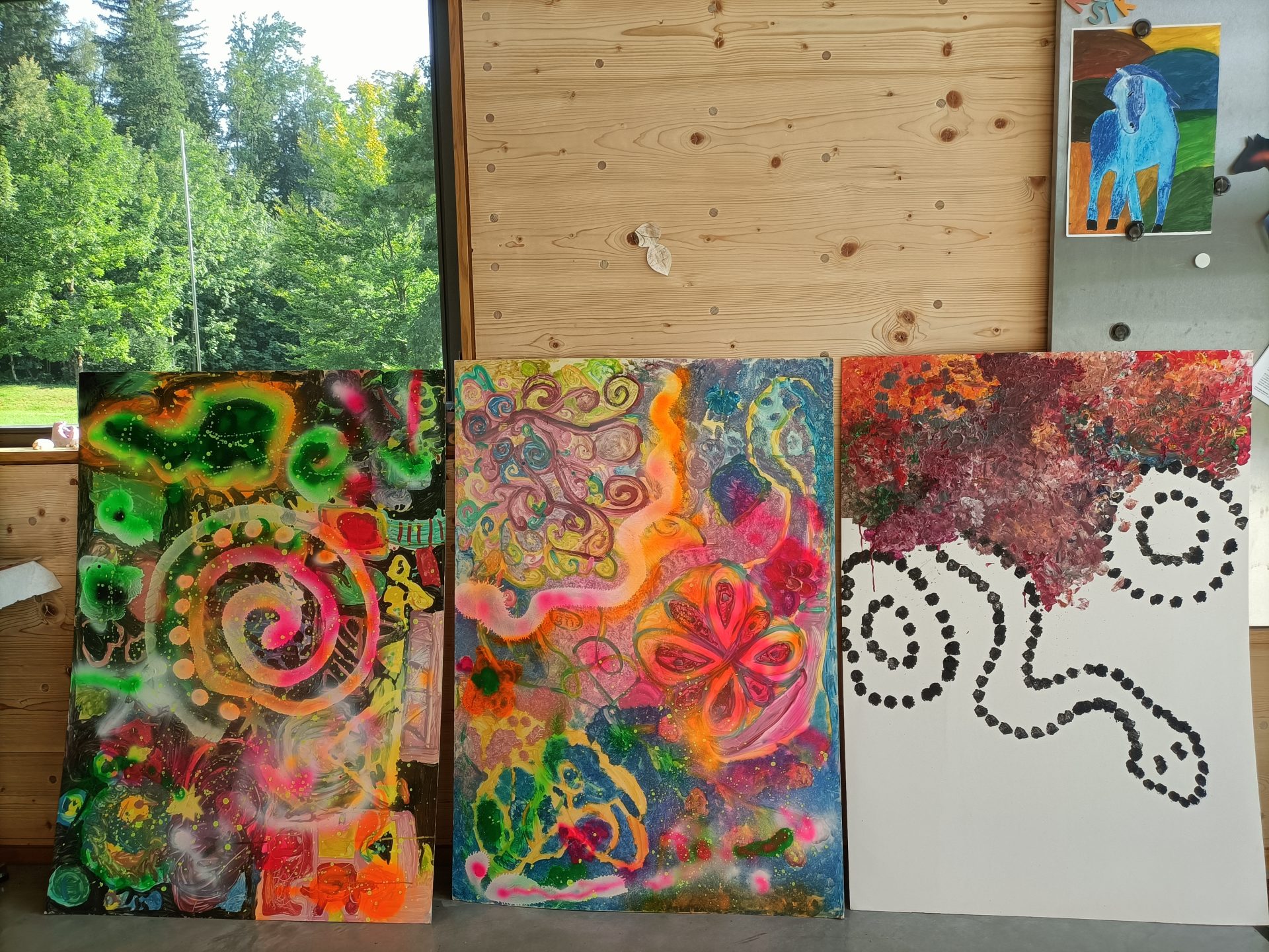 Drei farbenfrohe abstrakte Gemälde mit Wirbeln, Punkten und organischen Formen lehnen an einer Holzwand. Die Kunstwerke sind lebendig, mit Grün-, Rosa-, Gelb- und Orangetönen. Draußen sind durch ein Fenster grüne Bäume zu sehen.