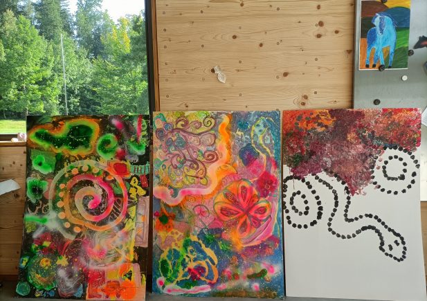 Drei farbenfrohe abstrakte Gemälde mit Wirbeln, Punkten und organischen Formen lehnen an einer Holzwand. Die Kunstwerke sind lebendig, mit Grün-, Rosa-, Gelb- und Orangetönen. Draußen sind durch ein Fenster grüne Bäume zu sehen.