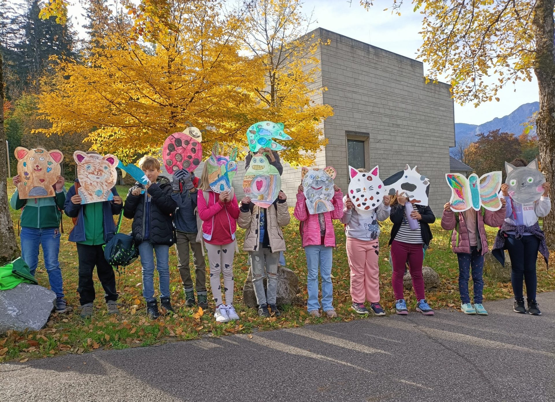 Eine Gruppe von Kindern steht im Herbst im Freien und hält sich große, bunte Tiermasken vor das Gesicht. Im Hintergrund sind Bäume mit gelben Blättern und ein modernes Gebäude zu sehen.