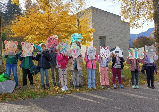 Eine Gruppe von Kindern steht im Herbst im Freien und hält sich große, bunte Tiermasken vor das Gesicht. Im Hintergrund sind Bäume mit gelben Blättern und ein modernes Gebäude zu sehen.