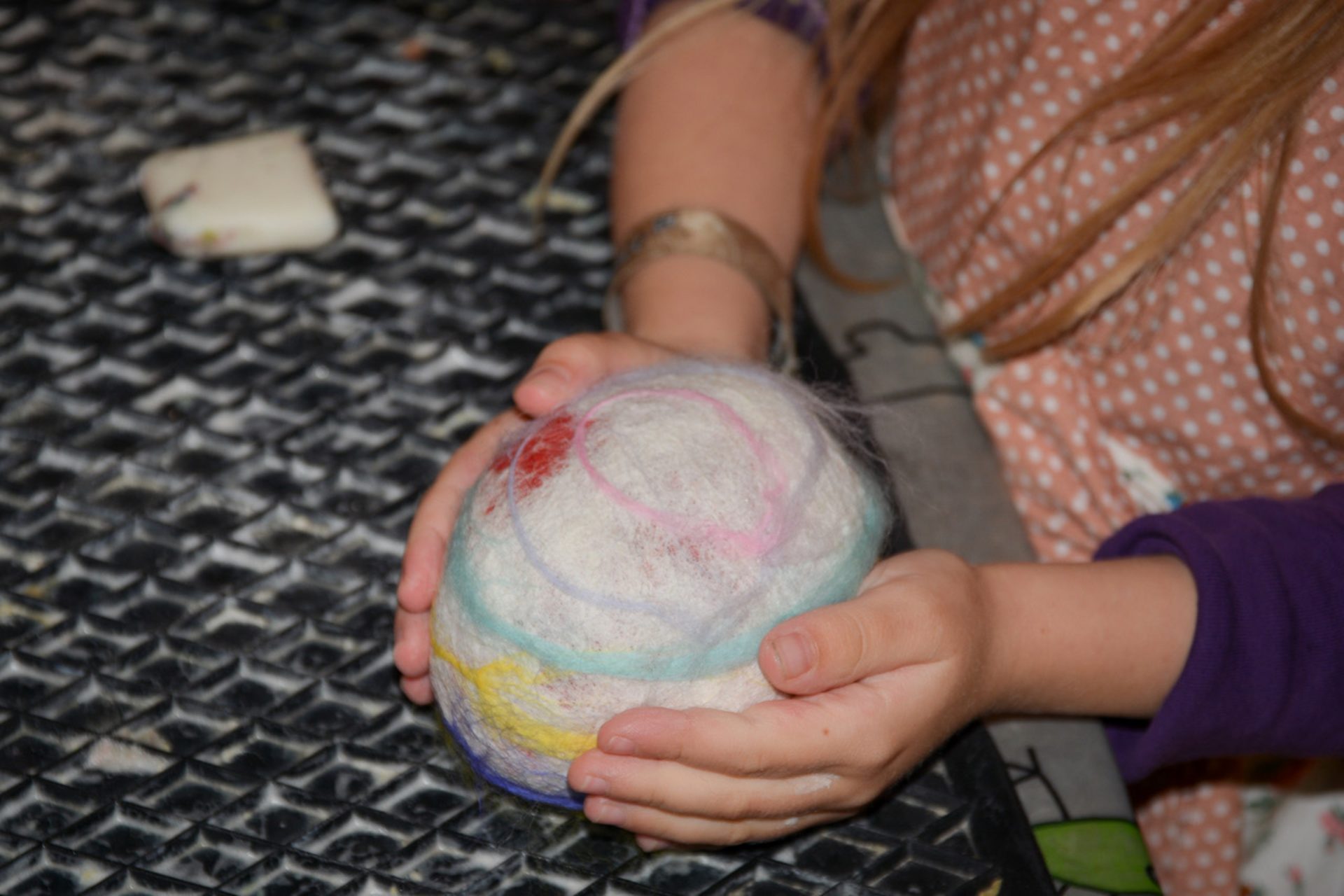 Ein Kind mit langem blondem Haar hält in beiden Händen ein rundes, bunt gefilztes Wollobjekt über einer strukturierten schwarzen Oberfläche. Ein kleiner rechteckiger Schwamm liegt daneben.