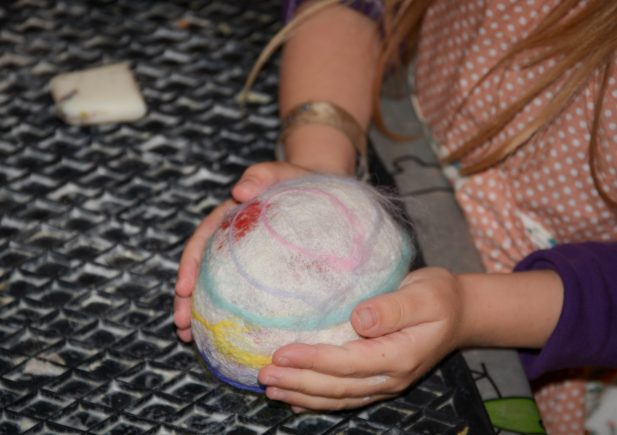 Ein Kind mit langem blondem Haar hält in beiden Händen ein rundes, bunt gefilztes Wollobjekt über einer strukturierten schwarzen Oberfläche. Ein kleiner rechteckiger Schwamm liegt daneben.