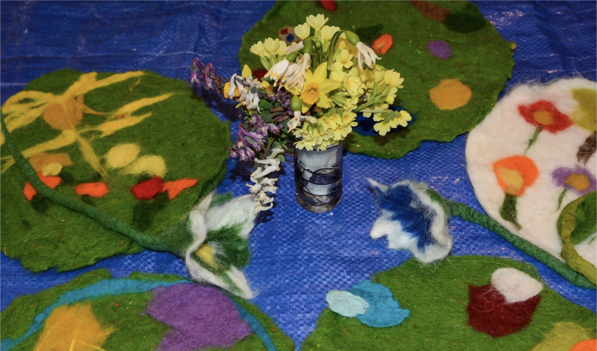 Eine kleine Glasvase mit gelben und weißen Wildblumen steht auf einer blauen Fläche, umgeben von bunten, handgemachten Filzmatten mit floralen und abstrakten Motiven.