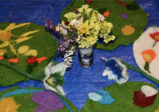 Eine kleine Glasvase mit gelben und weißen Wildblumen steht auf einer blauen Fläche, umgeben von bunten, handgemachten Filzmatten mit floralen und abstrakten Motiven.