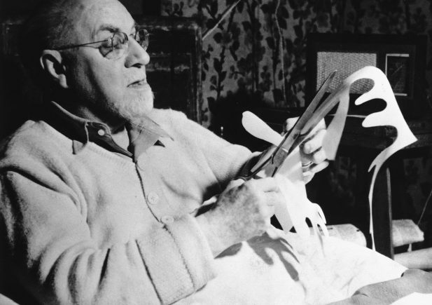 Ein älterer Mann mit Brille sitzt im Bett, hält eine Schere in der Hand und schneidet Formen aus weißem Papier aus. Er scheint auf seine Arbeit konzentriert zu sein, umgeben von Haushaltsgegenständen und einem gemusterten Vorhang im Hintergrund.