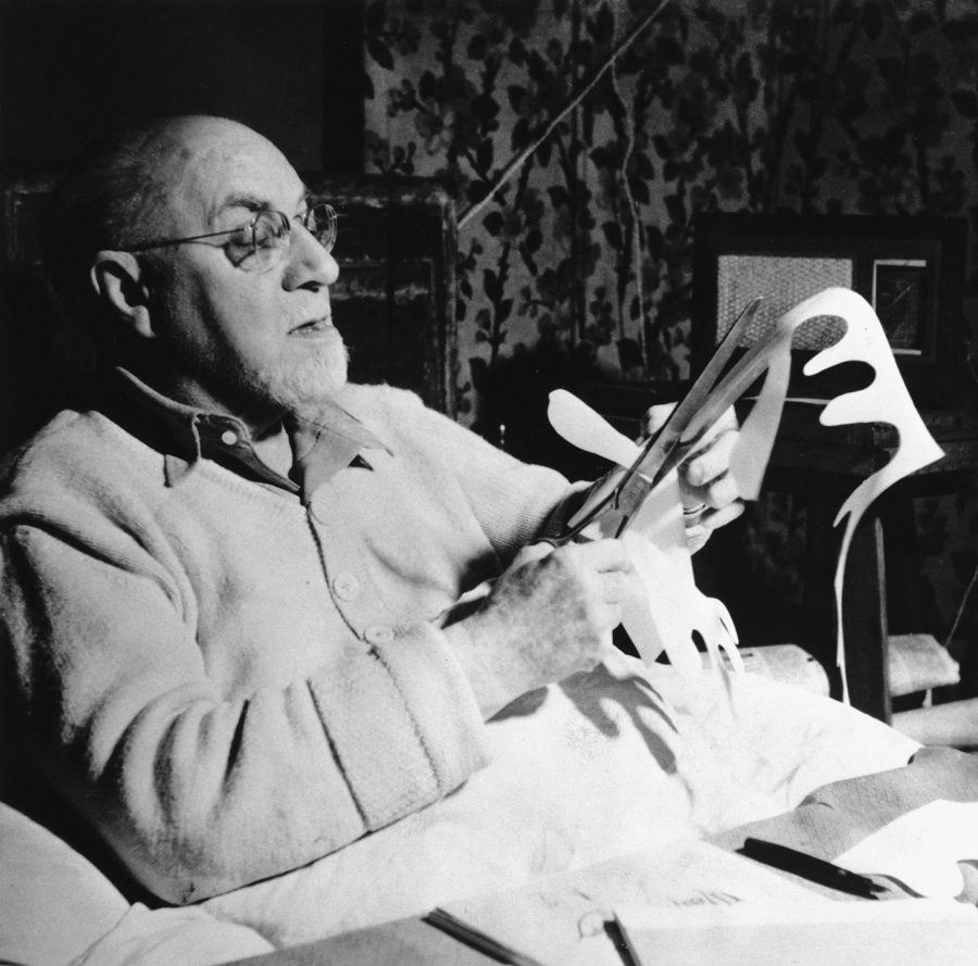 Ein älterer Mann mit Brille und Bart sitzt im Bett, hält eine Schere in der Hand und schneidet Formen aus weißem Papier aus. Im Hintergrund sind gemusterte Vorhänge und ein schwach beleuchtetes Zimmer zu sehen.