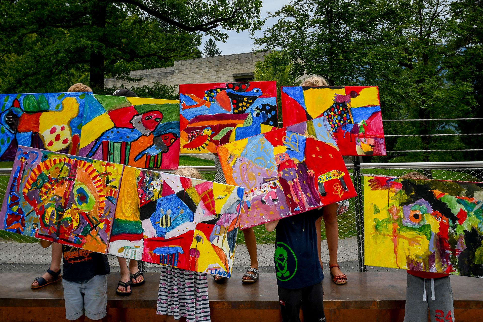 Kinder halten große, farbenfrohe abstrakte Gemälde hoch, die ihre Gesichter verdecken, und stehen im Freien mit Bäumen und einem Gebäude im Hintergrund. Die Kunstwerke sind lebendig und weisen verschiedene Formen und Muster auf.