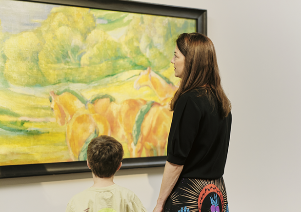 Eine Frau und ein Kind stehen vor einem farbenfrohen Gemälde mit Pferden und Grünpflanzen in einer Kunstgalerie und betrachten das Kunstwerk gemeinsam.