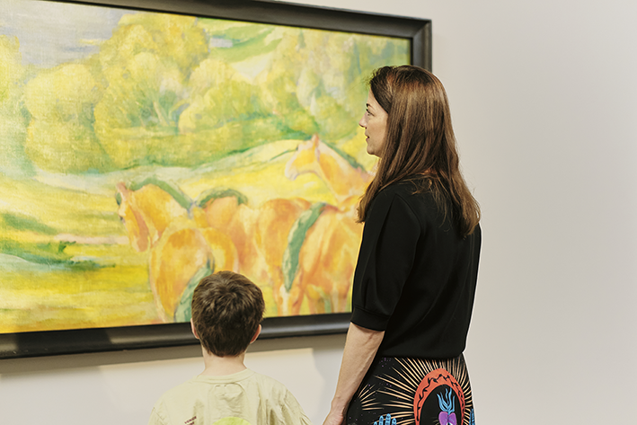 Eine Frau und ein Kind stehen vor einem farbenfrohen Gemälde mit Pferden und Grünpflanzen in einer Kunstgalerie und betrachten das Kunstwerk gemeinsam.