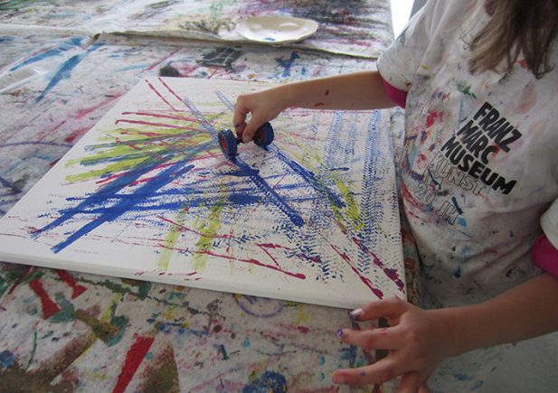 Ein Kind, das ein "Franz Marc Museum"-Shirt trägt, benutzt einen blauen Farbroller, um bunte, sich kreuzende Linien auf eine weiße Leinwand zu malen. Der Tisch und die Umgebung sind mit Spritzern und Abdrücken früherer Kunstwerke bedeckt.