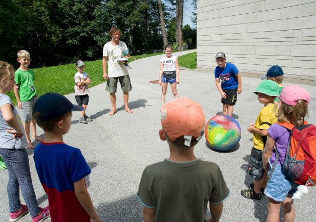 Eine Gruppe von Kindern steht an einem sonnigen Tag in einem Kreis, während ein Mann ein Klemmbrett hält. Ein Kind rollt einen bunten Ball in die Mitte, und alle scheinen mit einer Gruppenaktivität beschäftigt zu sein. Im Hintergrund sind Bäume und ein Gebäude zu sehen.