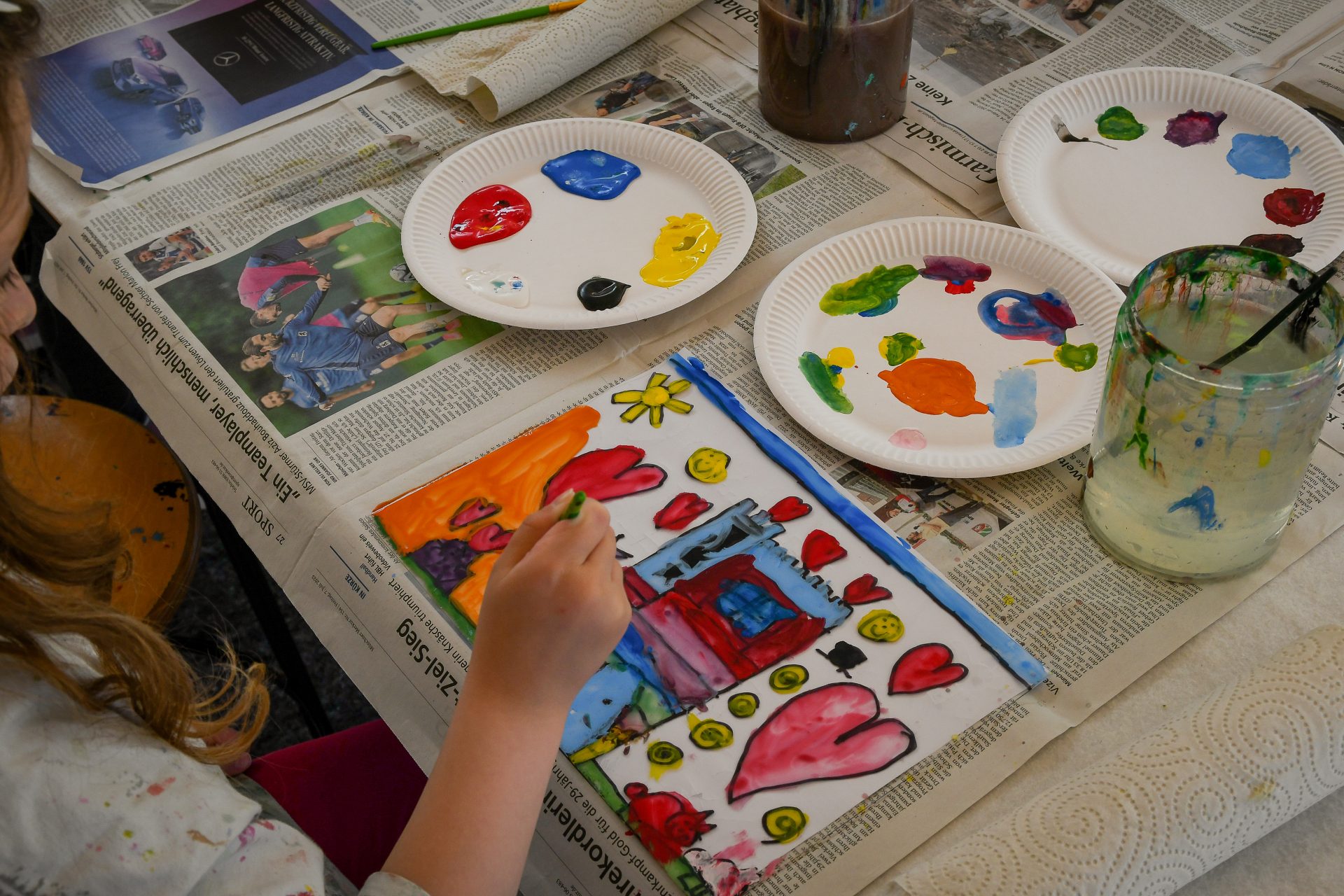 Ein Kind malt ein buntes Bild mit Herzen, einem Haus und einer Sonne auf Papier, umgeben von Tellern mit bunten Farbtupfern, Pinseln in einem Glas und Zeitungen, die den Tisch bedecken.