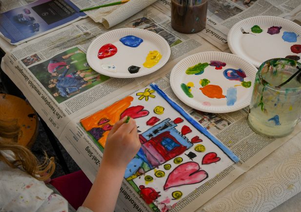Ein Kind malt ein buntes Bild mit Herzen, einem Haus und einer Sonne auf Papier, umgeben von Tellern mit bunten Farbtupfern, Pinseln in einem Glas und Zeitungen, die den Tisch bedecken.