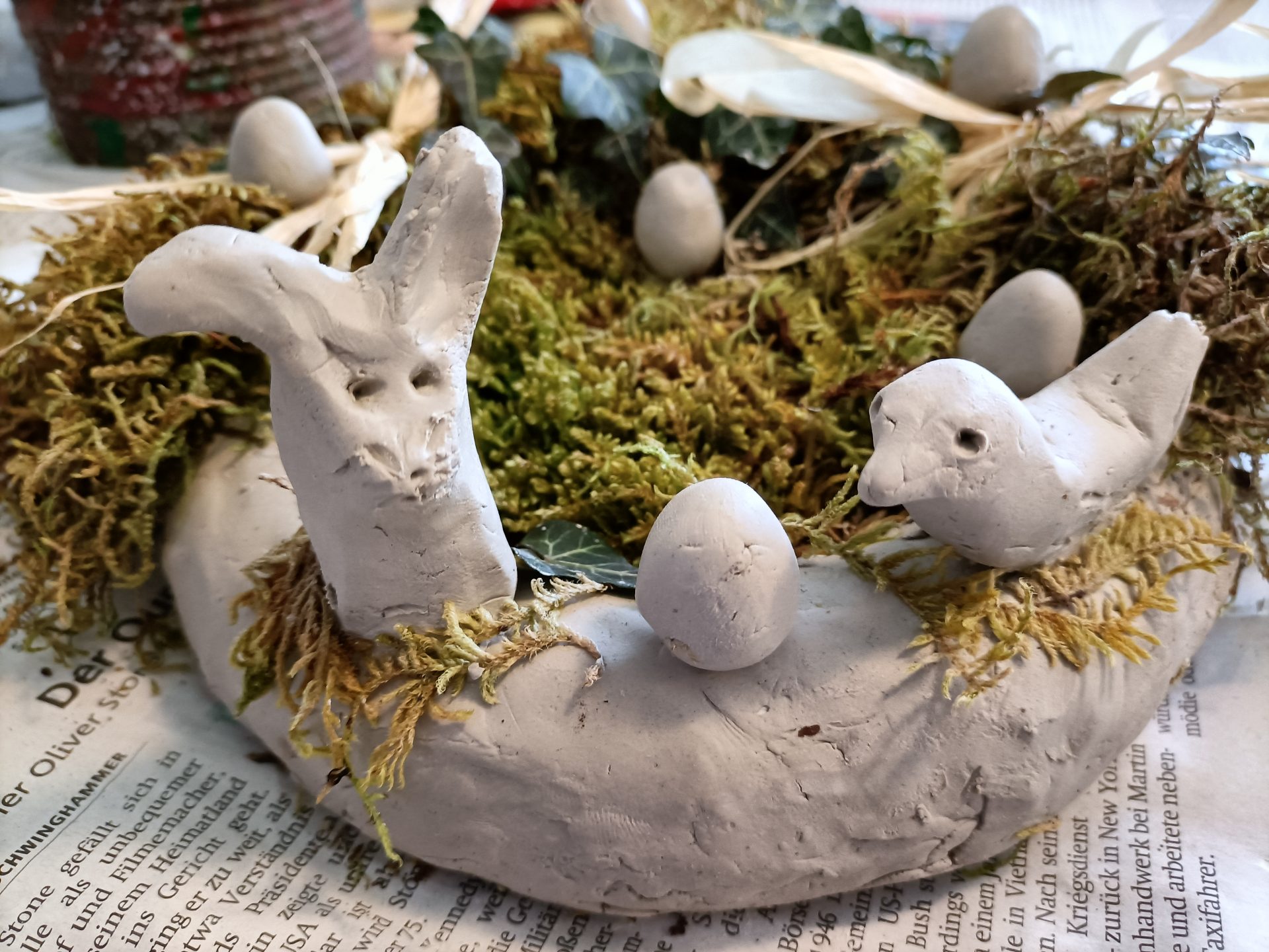 Ein handgefertigter Kranz aus Ton, verziert mit Moos, Toneiern, einem Tonhasen und einem Tonvogel, ausgestellt auf einer Zeitung.
