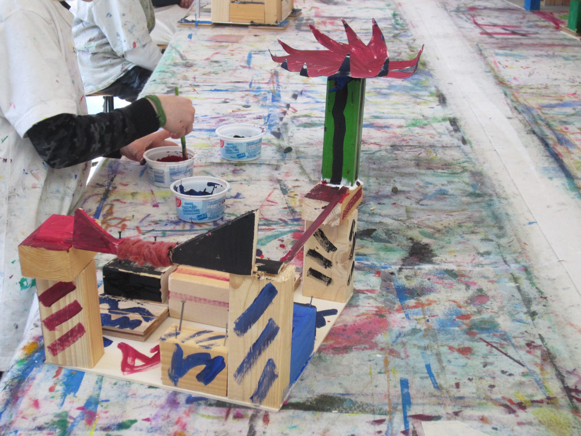 Eine Person bemalt eine bunte Holzskulptur mit abstrakten Formen auf einem Tisch, der mit Farbspritzern und -schlieren bedeckt ist und eine künstlerische und kreative Atmosphäre schafft.