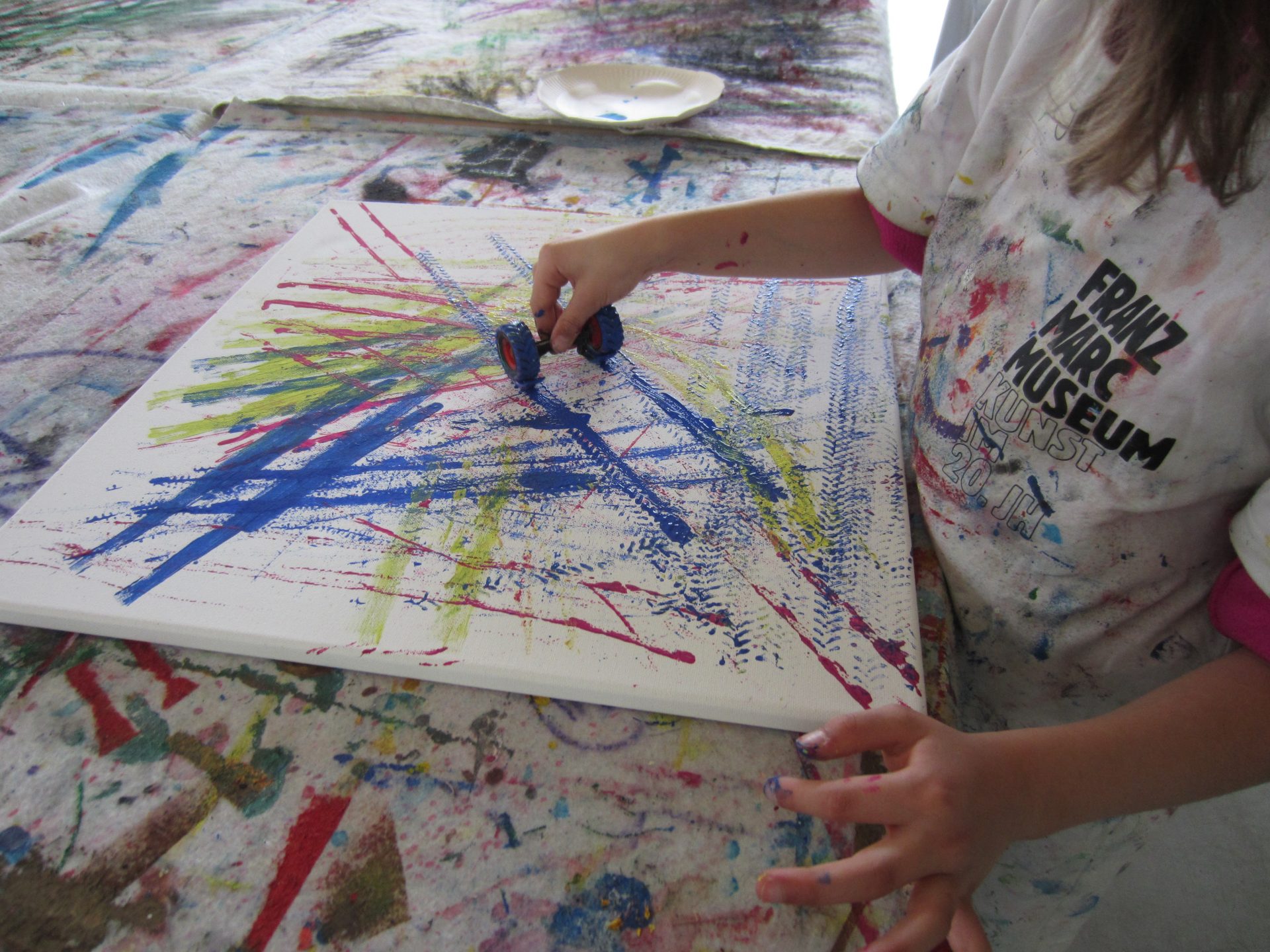 Ein Kind malt mit einem kleinen Roller blaue Linien auf eine weiße Leinwand und schafft so ein abstraktes Muster mit bunten, sich kreuzenden Linien. Der Tisch und das Hemd des Kindes sind mit Farbspuren bedeckt.