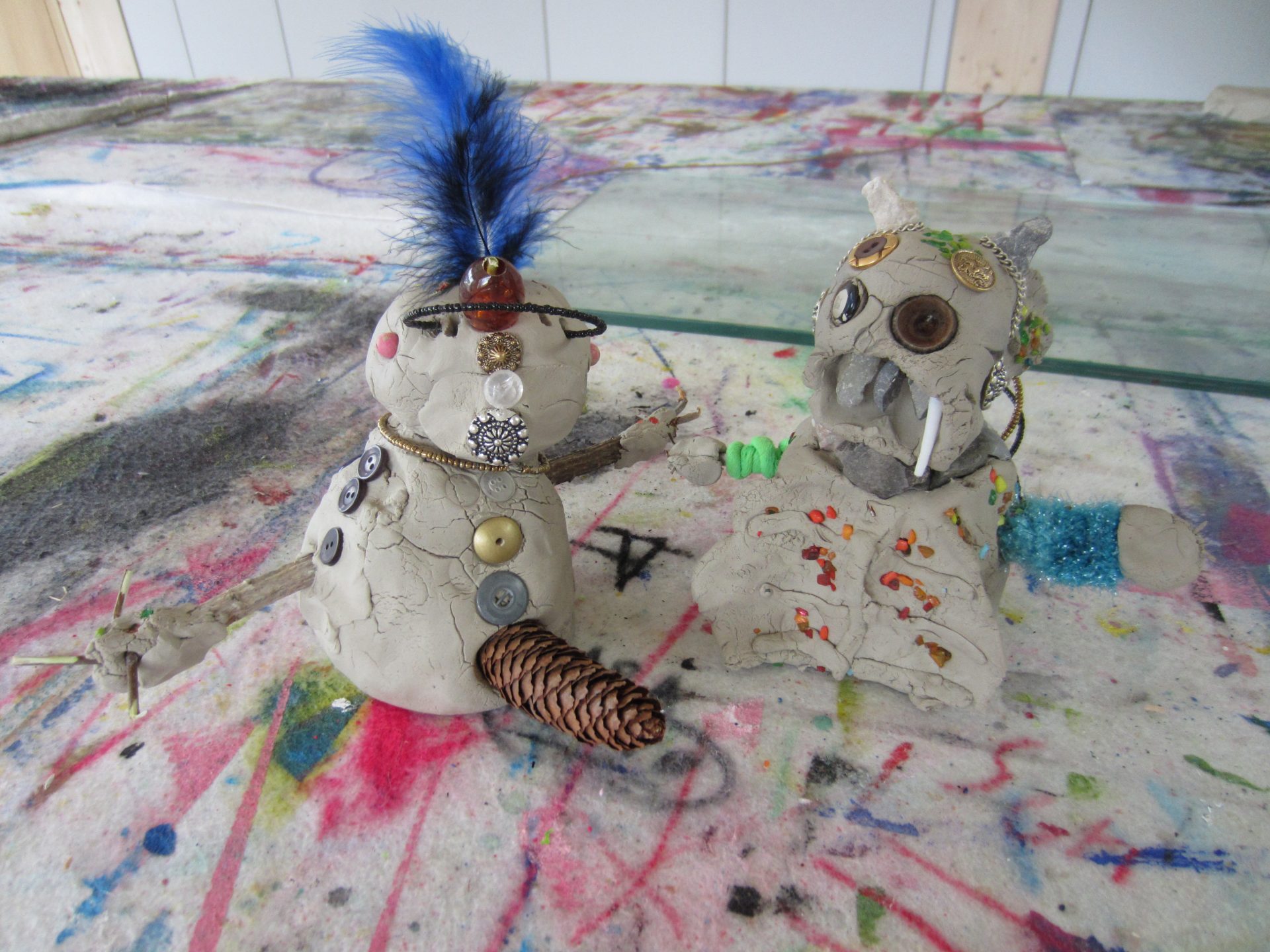 Zwei kleine Tonskulpturen auf einem bunten, mit Farbe bespritzten Tisch. Eine Figur hat eine blaue Feder, Knöpfe und Arme aus Kiefernzapfen; die andere hat Knöpfe, Stoßzähne und Muschelstücke, beide mit verschiedenen Fundstücken verziert.