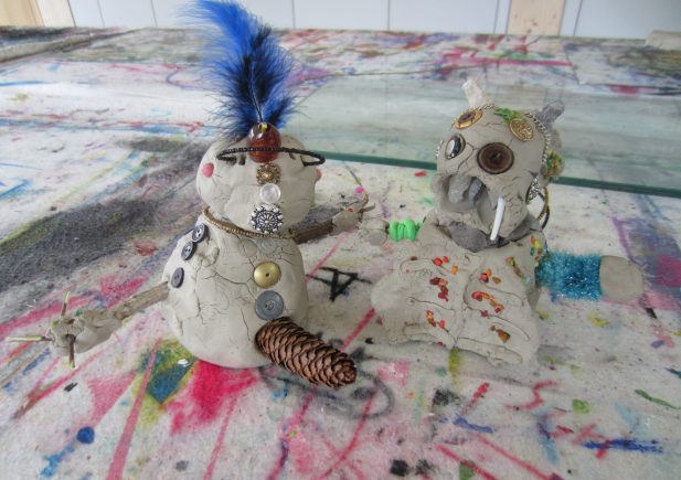 Zwei kleine Tonskulpturen auf einem bunten, mit Farbe bespritzten Tisch. Eine Figur hat eine blaue Feder, Knöpfe und Arme aus Kiefernzapfen; die andere hat Knöpfe, Stoßzähne und Muschelstücke, beide mit verschiedenen Fundstücken verziert.