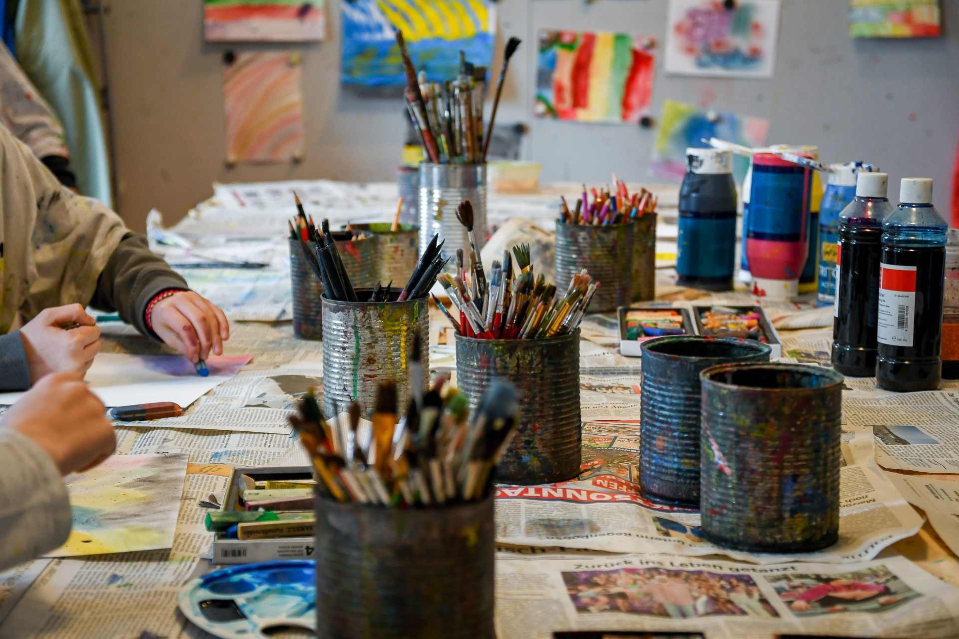 Ein Tisch, bedeckt mit Zeitungen, Pinseln in Blechdosen, Farbflaschen und Kunstzubehör. Zwei Personen zeichnen oder malen, und an der Wand im Hintergrund sind bunte Kunstwerke ausgestellt.