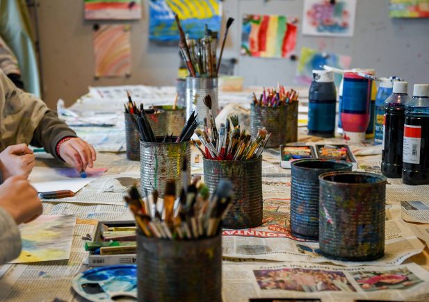 Ein Tisch, bedeckt mit Zeitungen, Pinseln in Blechdosen, Farbflaschen und Kunstzubehör. Zwei Personen zeichnen oder malen, und an der Wand im Hintergrund sind bunte Kunstwerke ausgestellt.