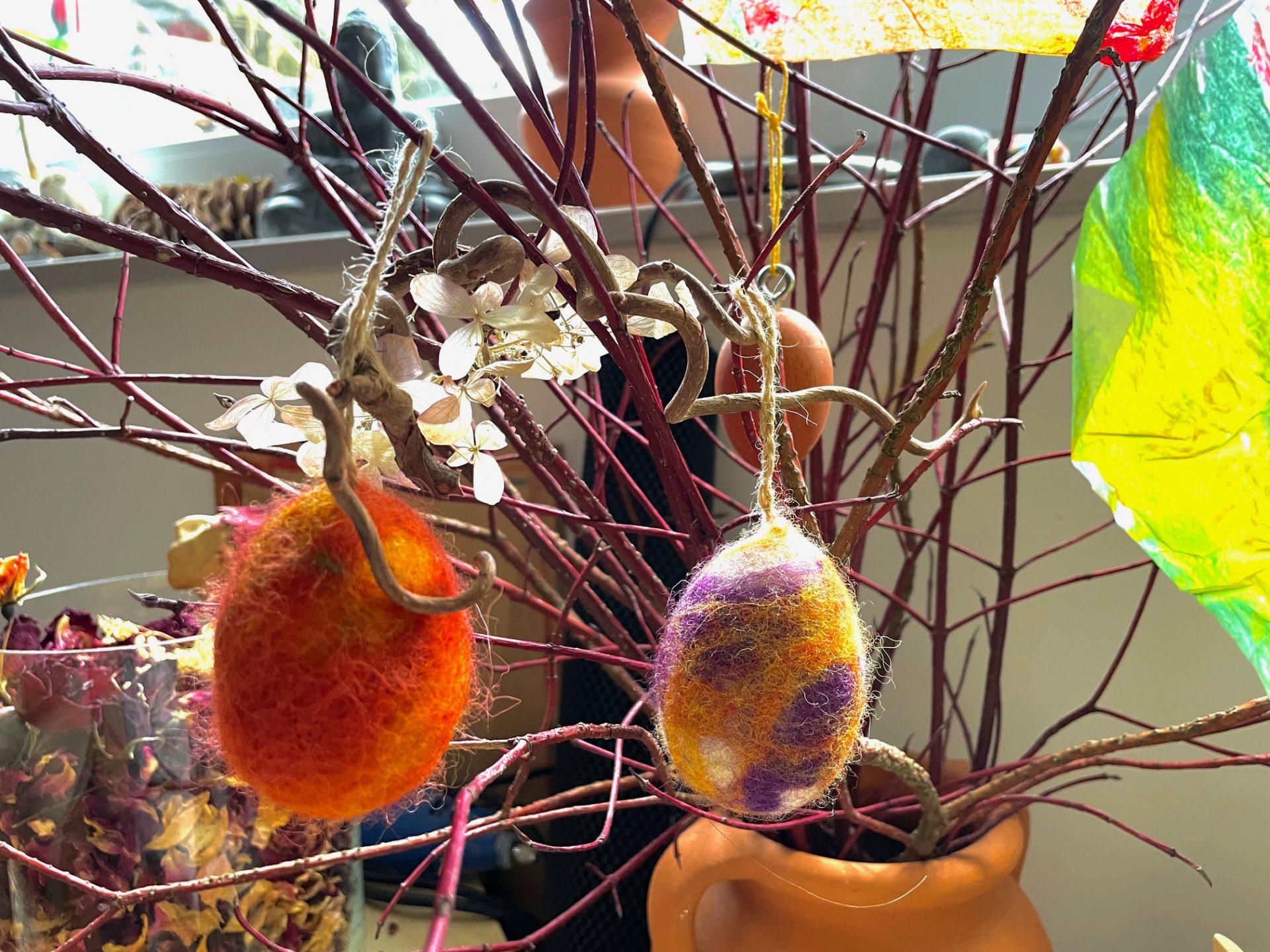 Zwei farbenfrohe Eierornamente aus Filz, eines orange-rot, das andere violett-gelb, hängen an kahlen Zweigen, die in einer Vase angeordnet sind. Getrocknete Blumen und buntes Seidenpapier schmücken den Hintergrund.