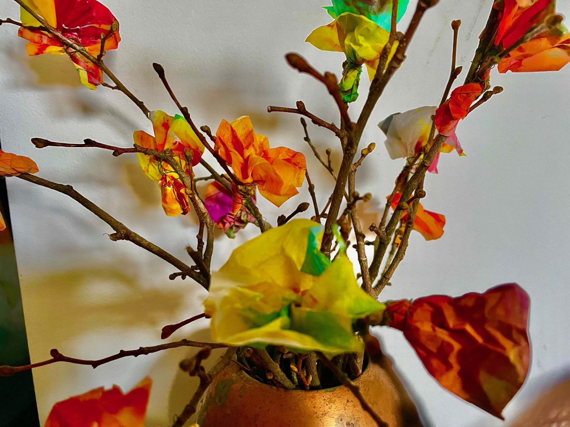 Die in einer Vase arrangierten Zweige sind mit bunten Seidenpapierblumen in Orange-, Gelb-, Grün- und Rottönen geschmückt, die vor einem unifarbenen Hintergrund ein fröhliches Bild ergeben.