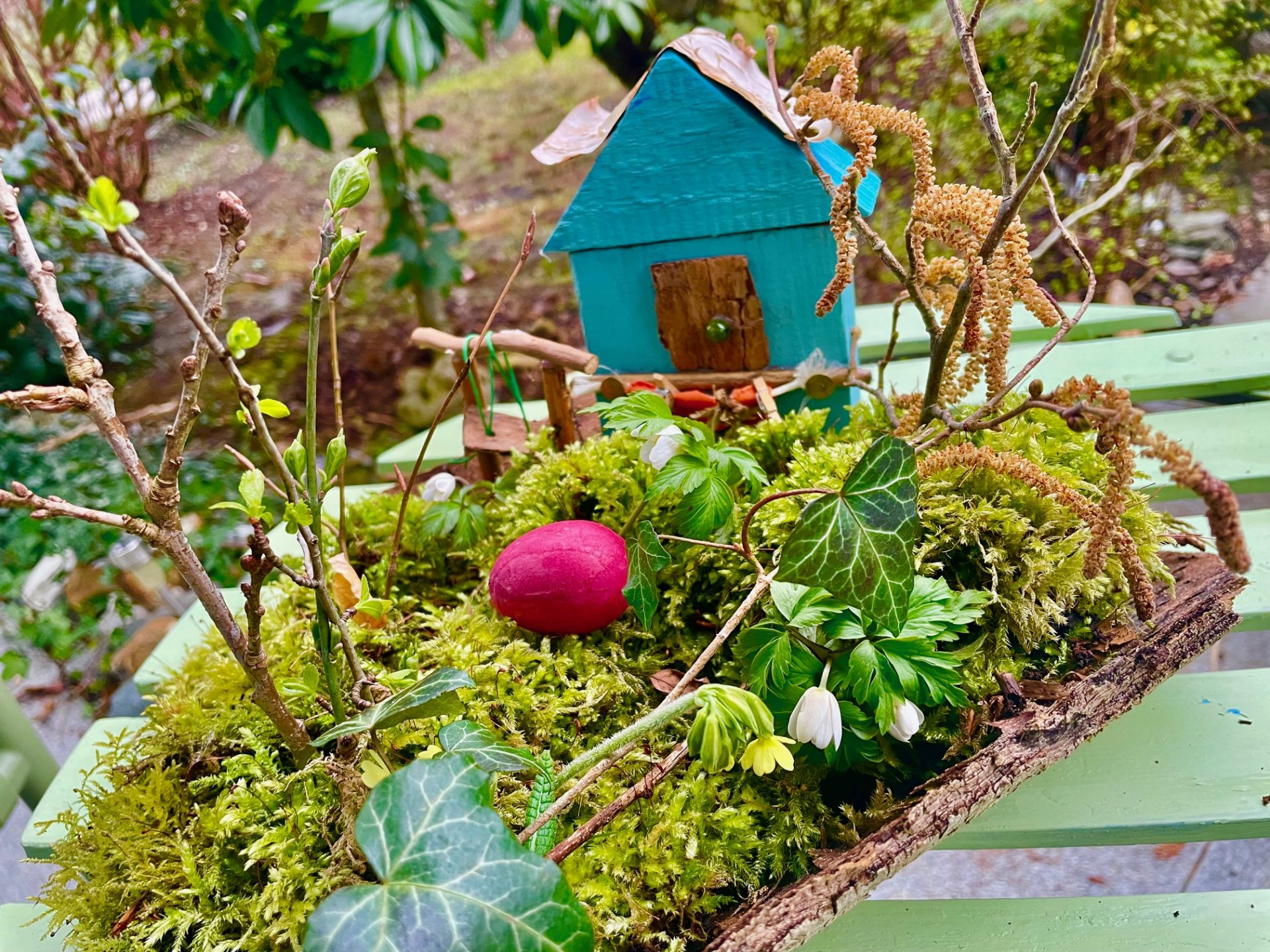 Ein kleines blaues Holzhaus steht auf einem grünen, moosbewachsenen Tablett, umgeben von Zweigen, Blättern, Blumen und einem einzelnen roten Ei. Die Szene befindet sich im Freien auf einer hellgrünen Holzfläche mit einem Gartenhintergrund.