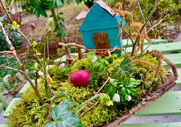 Ein kleines blaues Holzhaus steht auf einem grünen, moosbewachsenen Tablett, umgeben von Zweigen, Blättern, Blumen und einem einzelnen roten Ei. Die Szene befindet sich im Freien auf einer hellgrünen Holzfläche mit einem Gartenhintergrund.